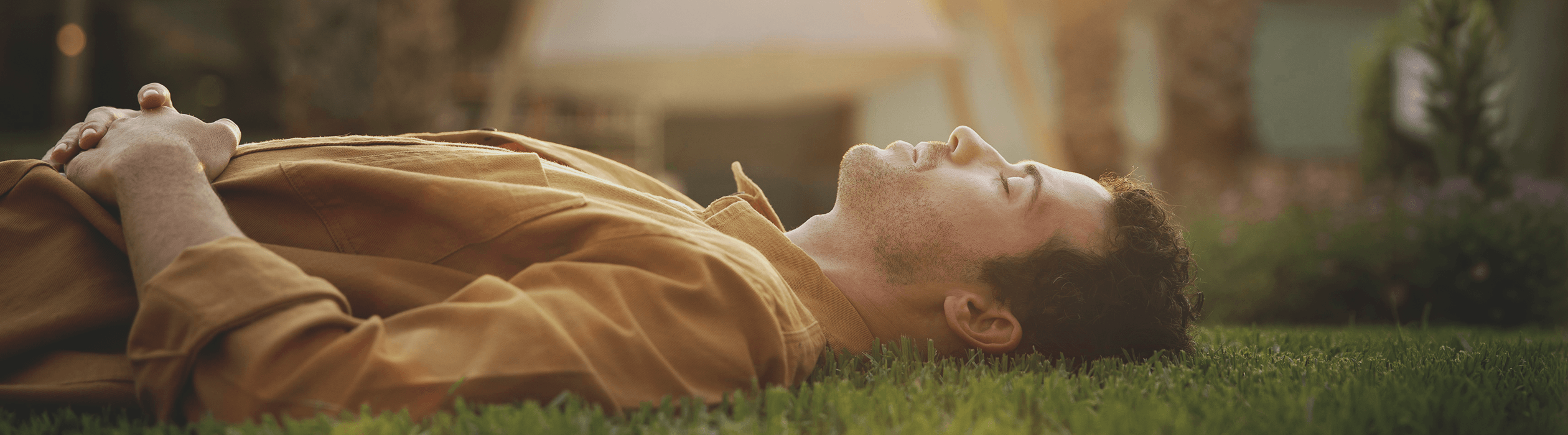 Man lying on his back on a grassy lawn with eyes closed, wearing a tan shirt and smiling faintly during a sunny day.